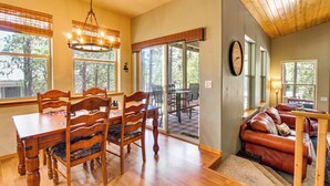 A dining area with a wooden table and chairs, a chandelier, and large windows with blinds. - Walk to Rainbow Lake: Peaceful Cabin w/ Fire Pit (Lakeside)