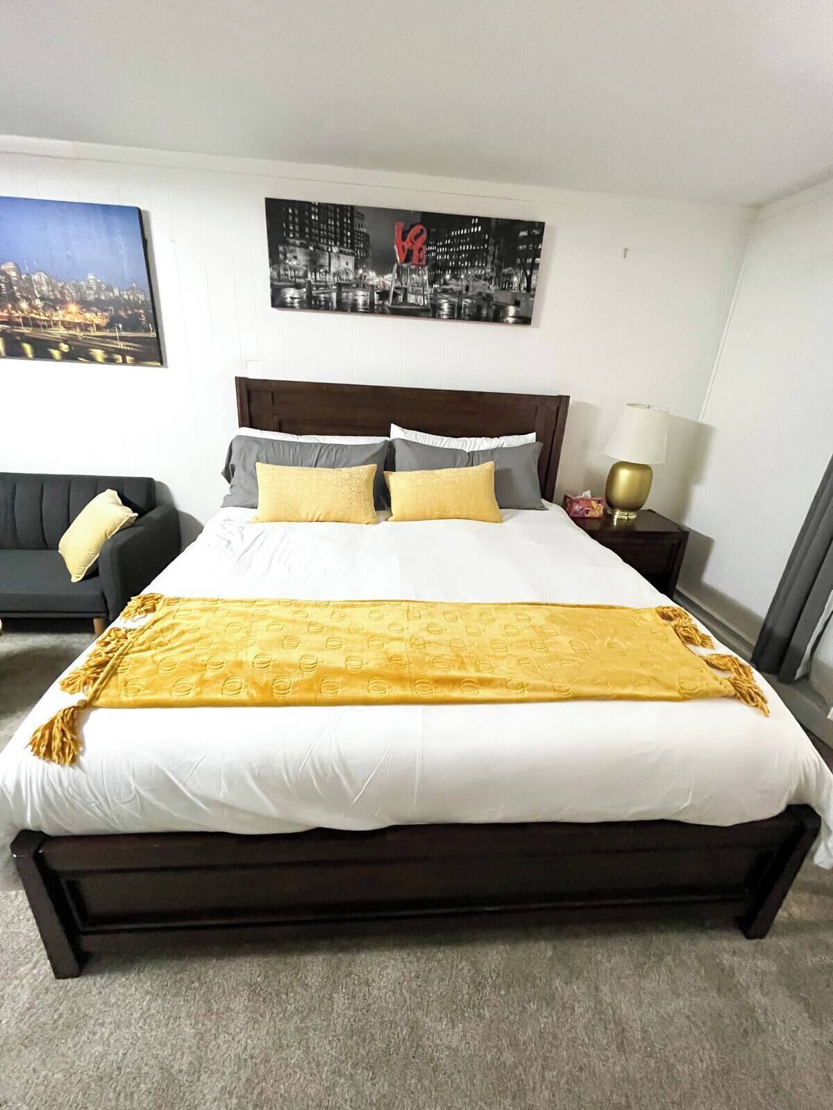 A hotel room with a large bed, a nightstand with a lamp, a couch, and two framed pictures on the wall.