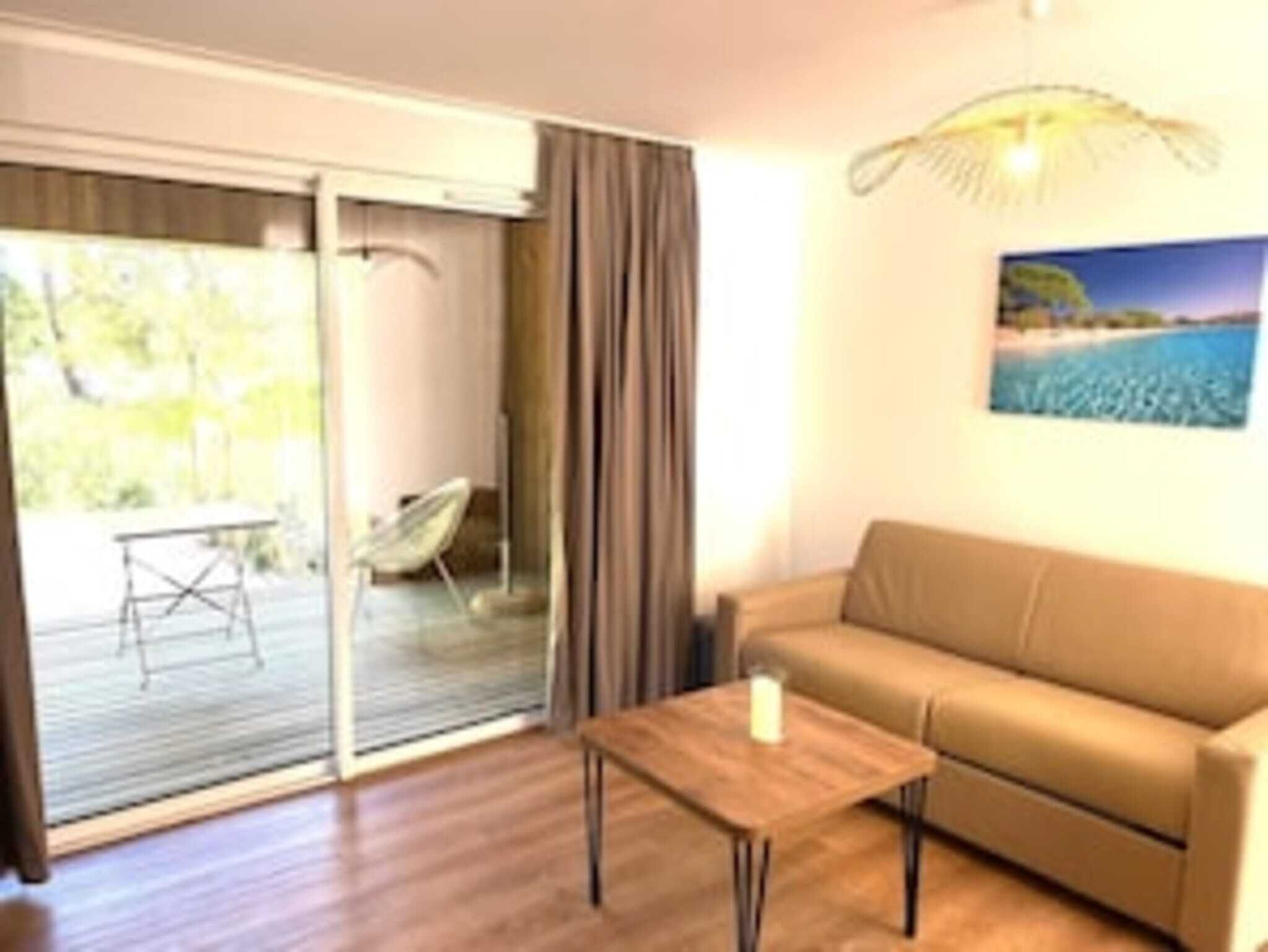 Ett rum med en soffa, ett litet bord och en stol. Det finns en skjutdörr i glas som leder ut till ett utomhusområde med ett bord och en stol. En tavla hänger på väggen.