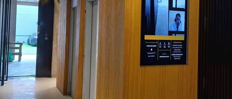 A hallway with wooden paneling and a security camera. There is a sign with contact information and a view of the outside.