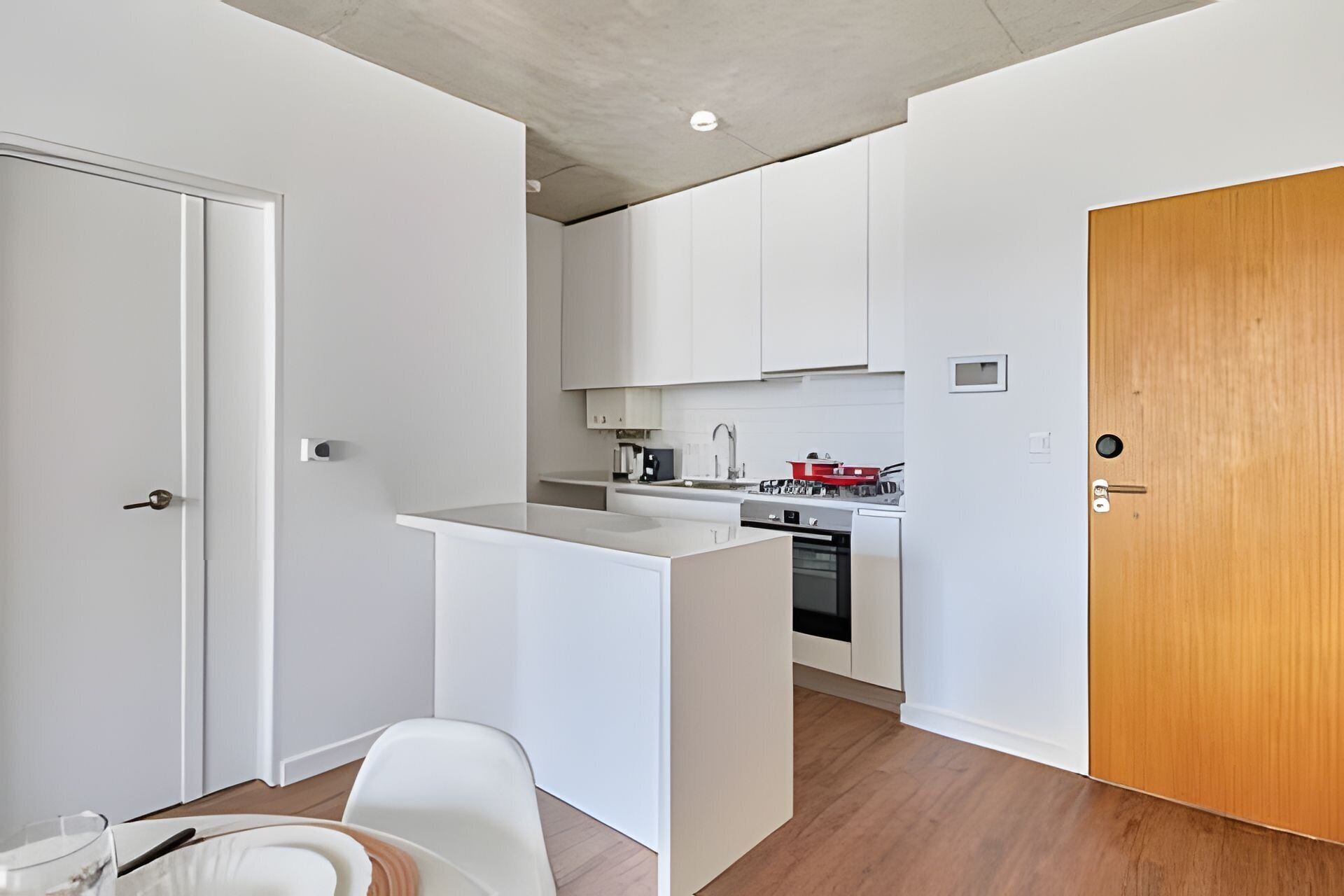 Una cucina compatta con mobili bianchi, una porta in legno e un piccolo spazio pranzo con una sedia e un tavolo bianchi.