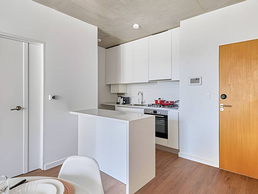 Una cucina compatta con mobili bianchi, una porta in legno e un piccolo spazio pranzo con una sedia e un tavolo bianchi.