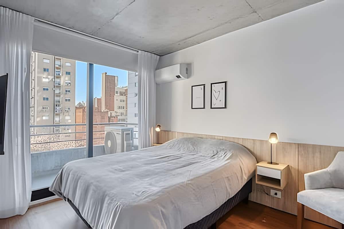 Een moderne slaapkamer met een groot bed, een houten hoofdbord, een nachtkastje, een stoel en uitzicht op gebouwen door een groot raam.