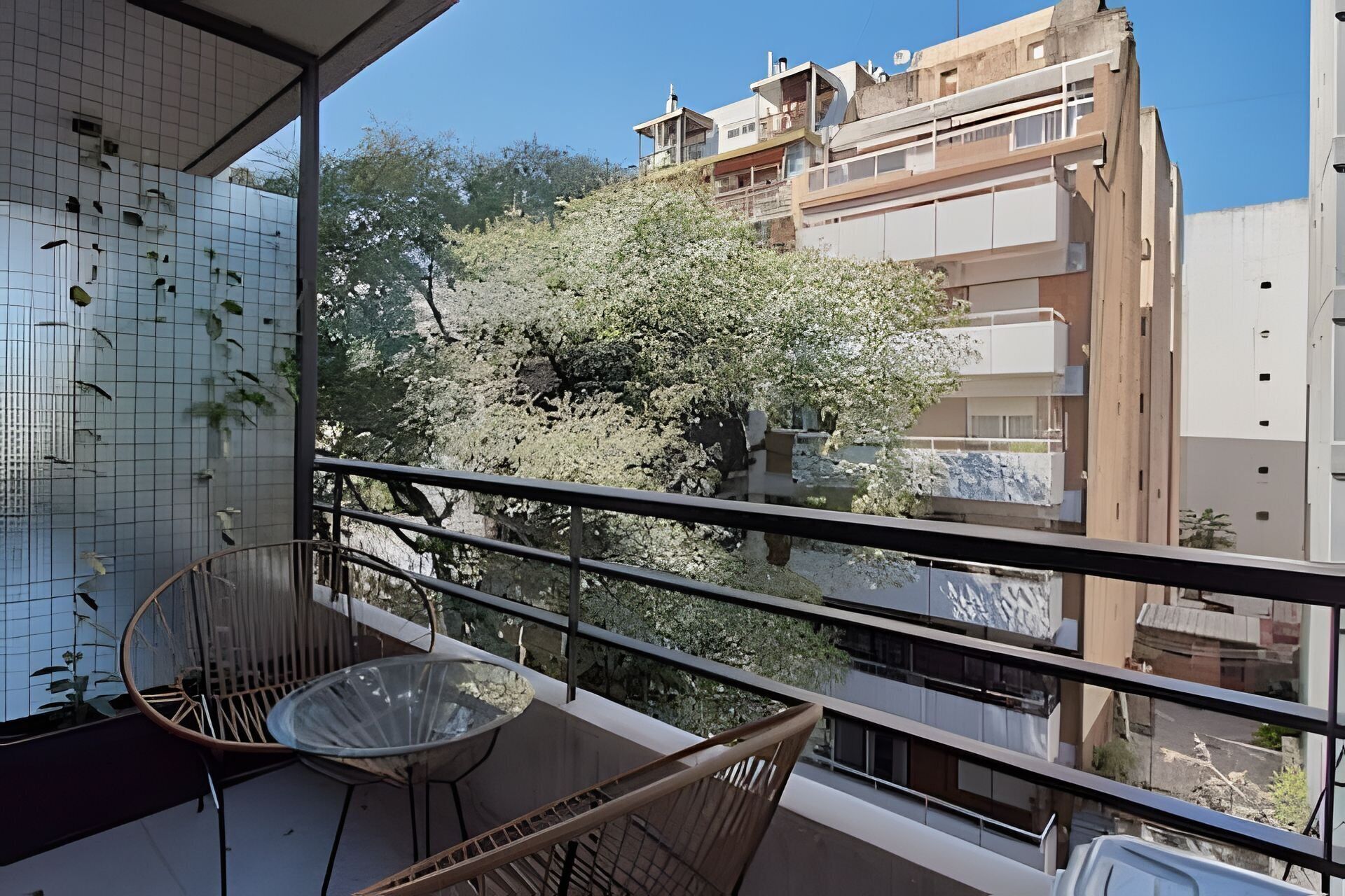 Ein Balkon mit Stuhl und Tisch, mit Blick auf eine Wohngegend mit Bäumen und Gebäuden.