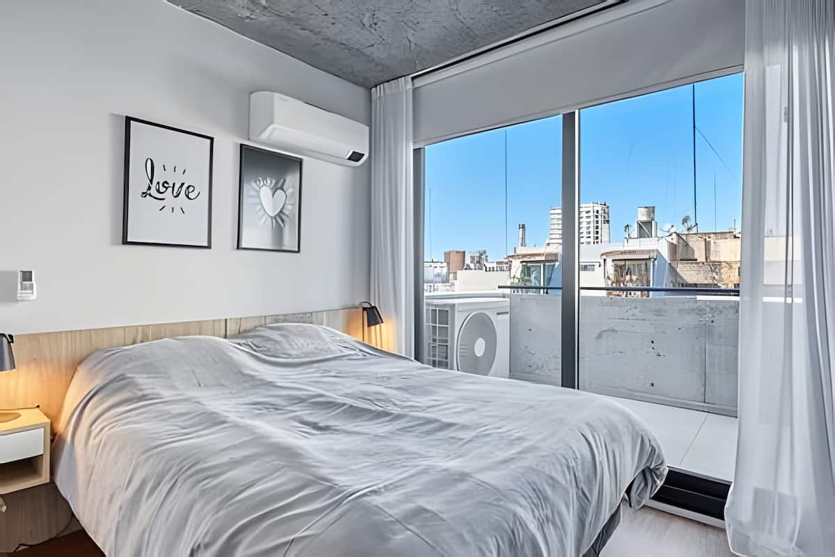 Een moderne slaapkamer met een groot bed, stadszicht door een raam en een plafondventilator.