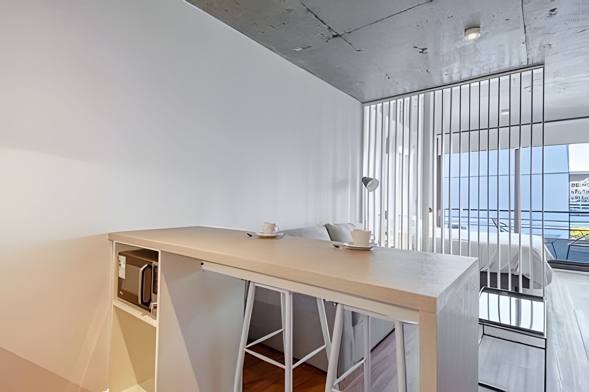 A modern, minimalist room with a long, white desk, a small seating area, and a large window with vertical blinds.