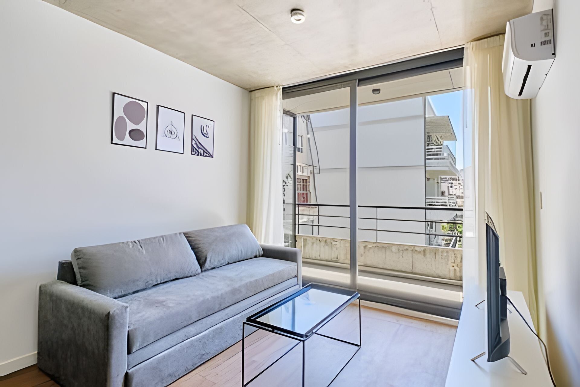Uma sala de estar moderna com um sofá cinza, uma mesa de centro de vidro e uma grande janela com vista para edifícios.