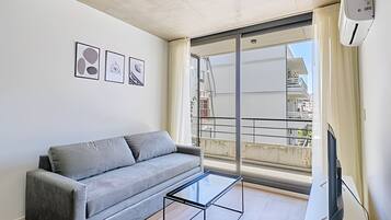 A modern living room with a grey sofa, a glass coffee table, and a large window with a view of buildings.