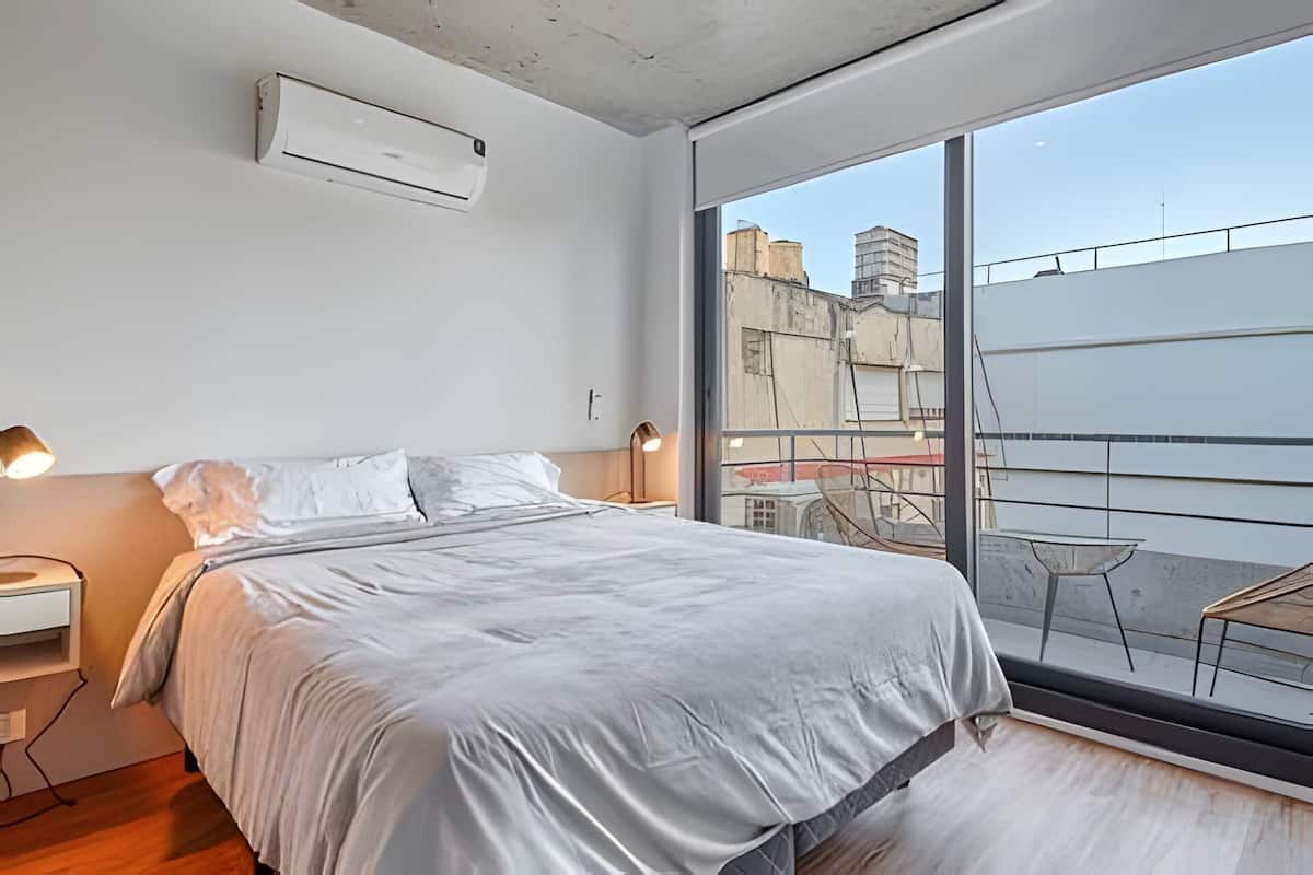 Een moderne slaapkamer met een groot bed, nachtlampjes en uitzicht op de stad vanaf een balkon.
