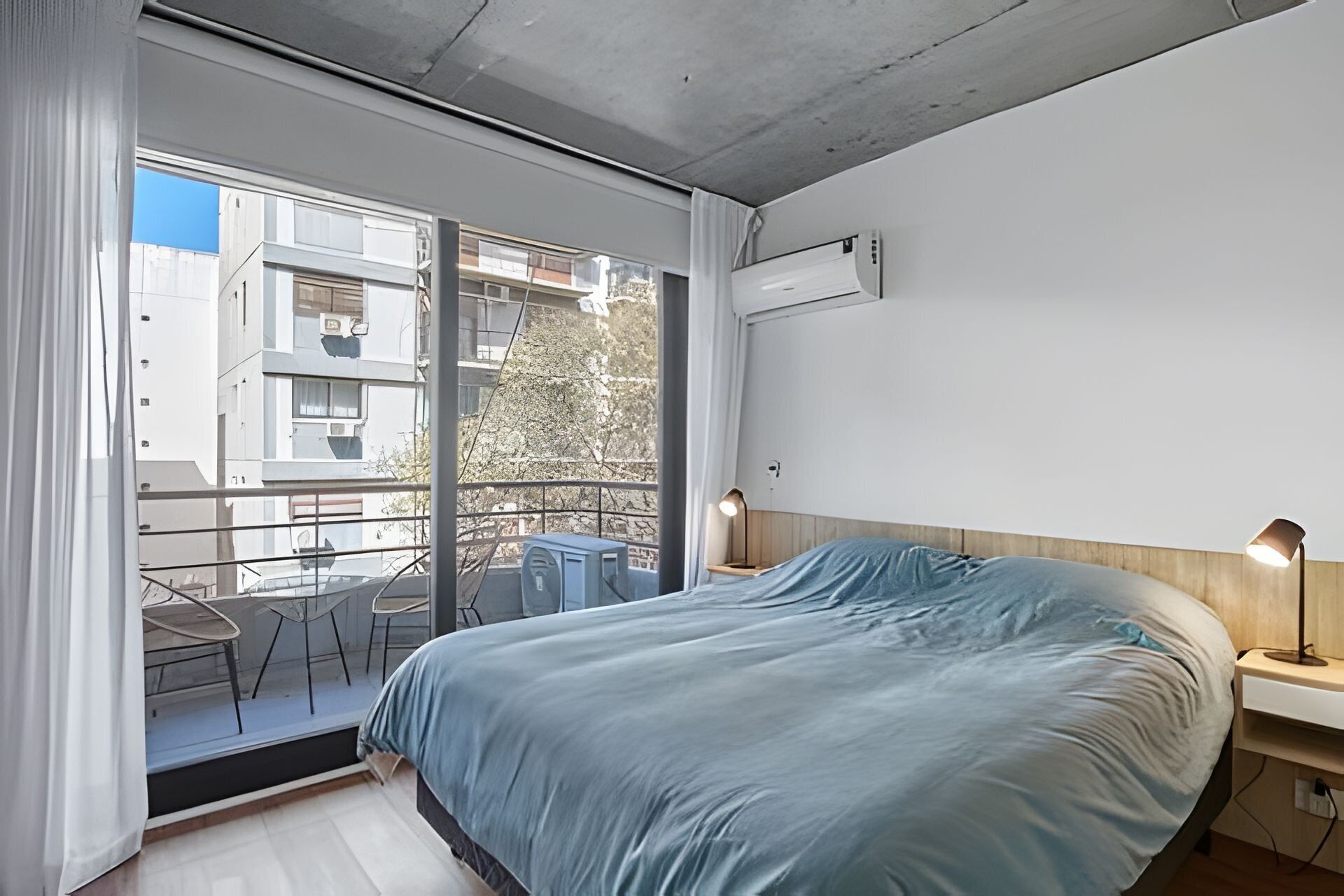 Una camera da letto con un letto, un comodino, una lampada e una finestra con vista su un balcone.