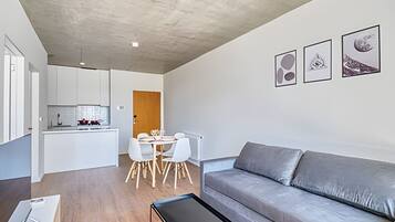 Un espacio de estar moderno con sofá gris, mesa de comedor blanca y zona de cocina.