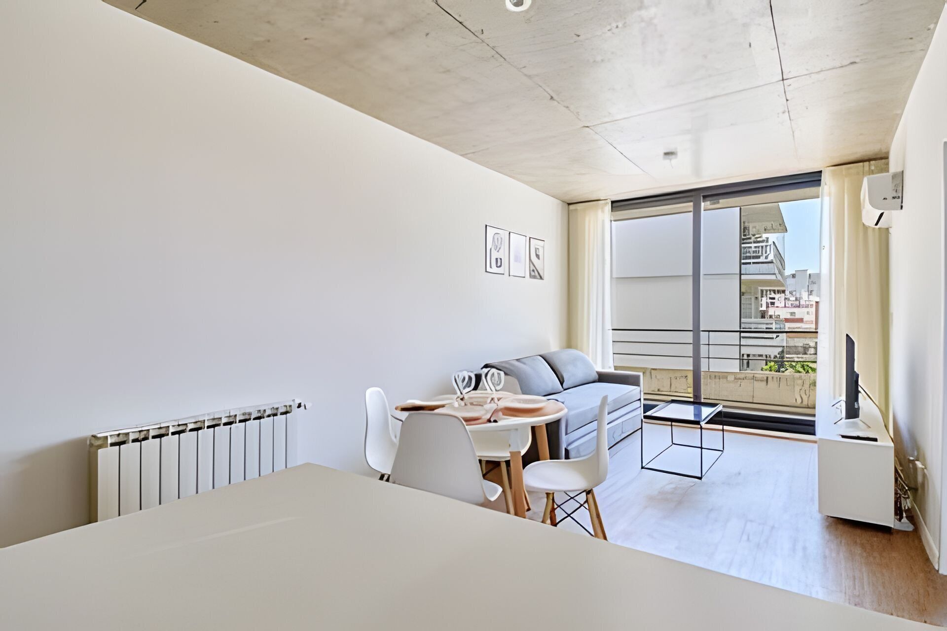 A modern, minimalist living room with a sofa, a dining table, and chairs. There is a large window providing a view of the city.