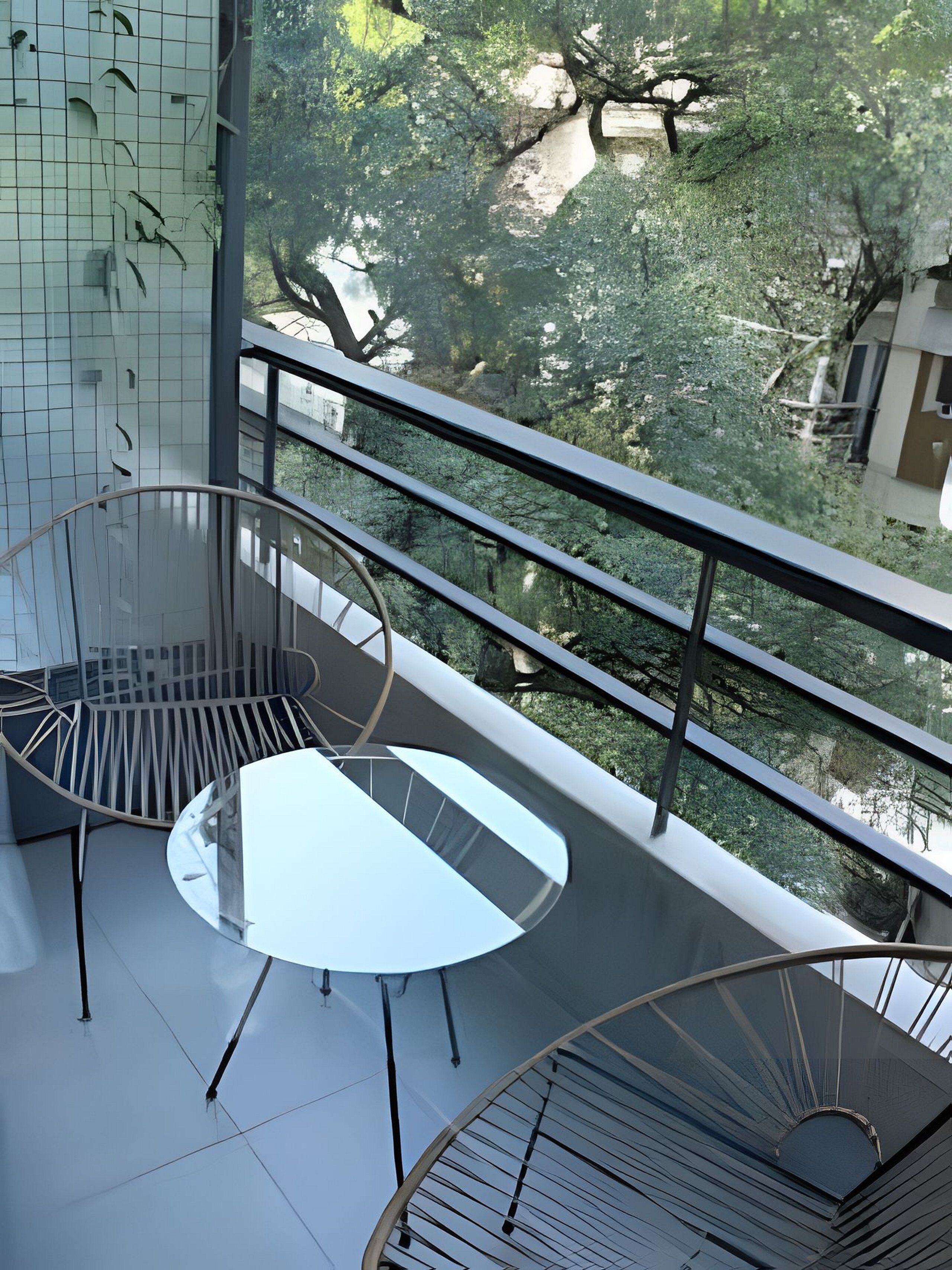 Un balcone moderno con un tavolo in vetro e sedie in metallo, affacciato su un rigoglioso giardino e una struttura in pietra.