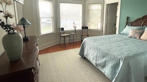 A bedroom with a large bed, a desk, and a chair. There are windows with blinds, a vase with flowers, and a ceiling fan. - Relax Inn The Grove (Forest Grove)
