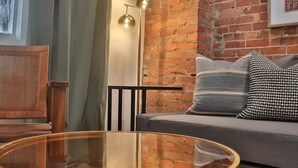 Ein gemütlicher Wohnbereich mit Backsteinwand, grauem Sofa mit gemusterten Kissen, rundem Glastisch und schwarzem Sessel.