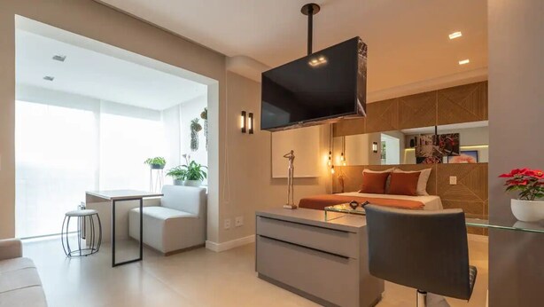 A modern living room with a sofa, a coffee table, a TV mounted on the wall, and a small dining area with a table and chairs. - Charming Studio - GRU 2001 (Jardim Flor da Montanha Guarulhos)