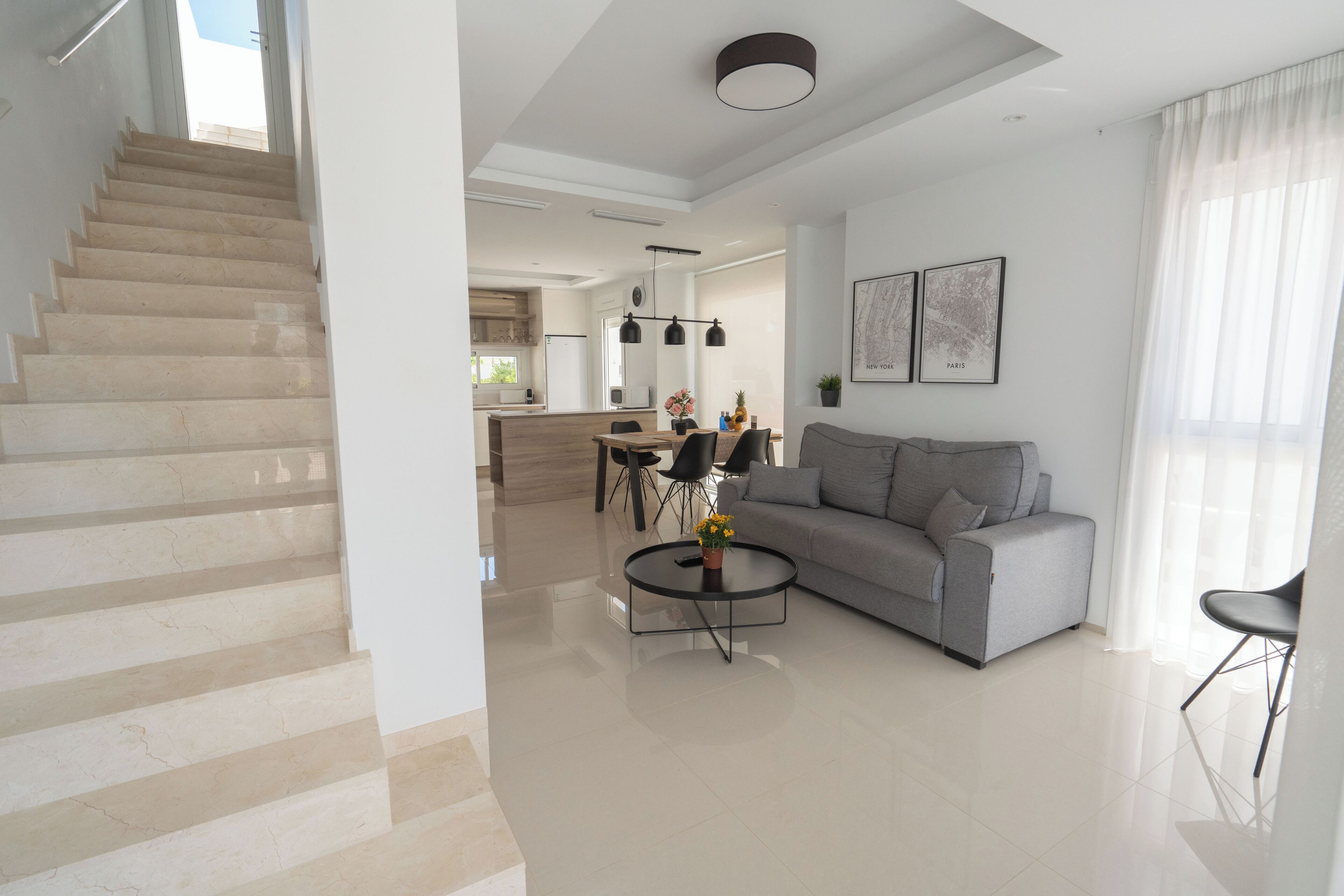 Una sala de estar moderna con una escalera, un sofá gris, una mesa de centro y un comedor con una cocina al fondo.
