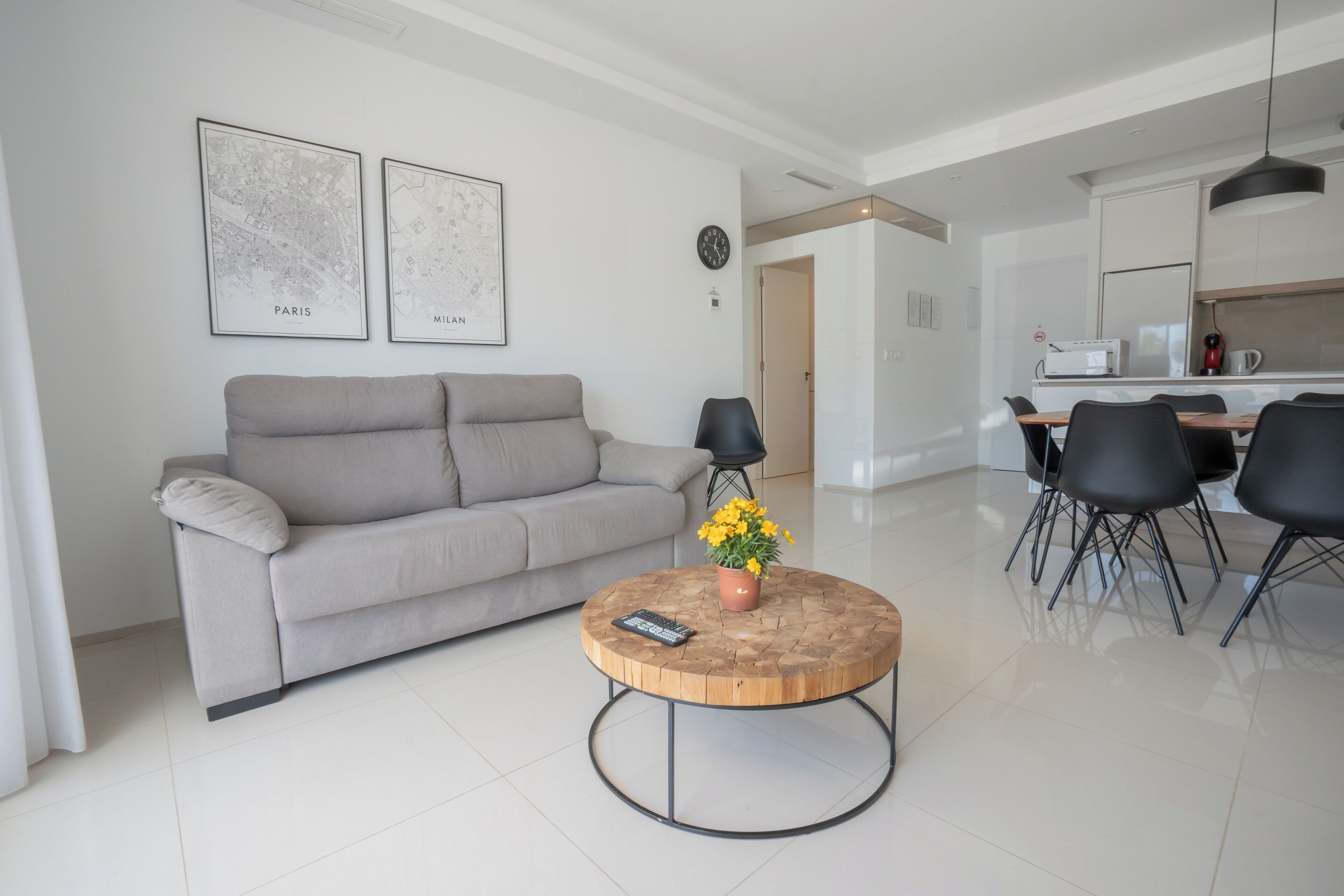 Una sala de estar moderna con un sofá gris, una mesa redonda de centro con un jarrón de flores y una zona de comedor con una mesa blanca y sillas negras.