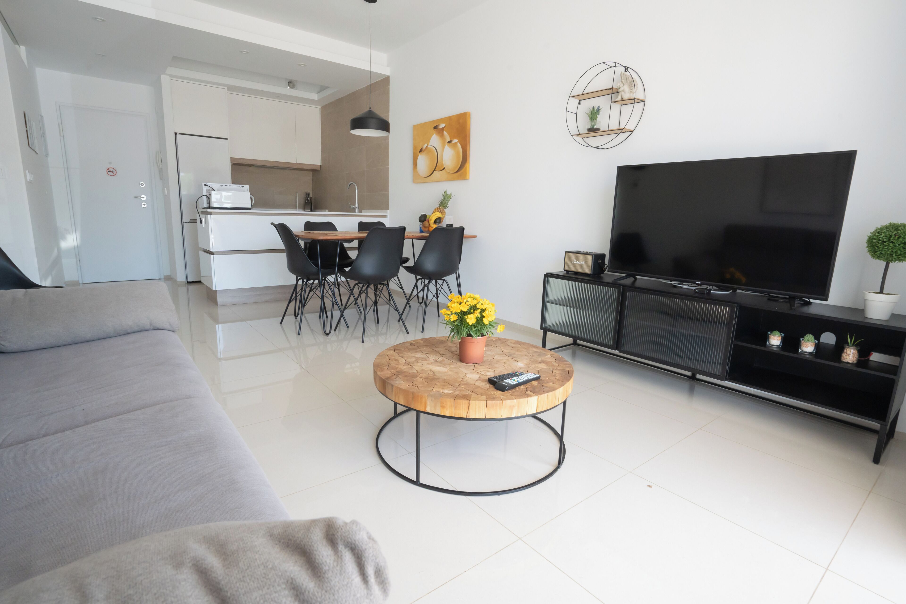 Een moderne woonkamer met een bank, een ronde salontafel met een bloevas, een eethoek met stoelen, een keuken op de achtergrond en een grote televisie op een zwart plateau.