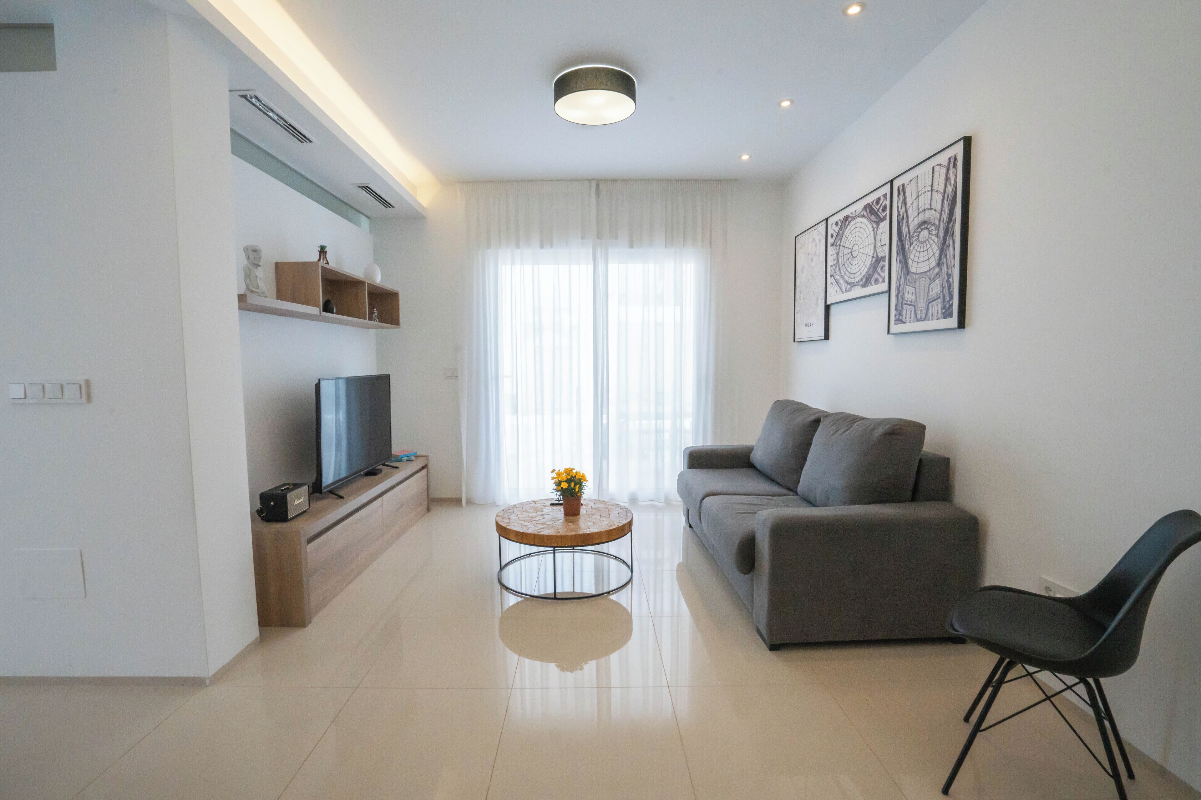 Una sala de estar moderna con un televisor de pantalla plana, un sofá gris, una mesa de centro redonda con un florero y una silla negra.