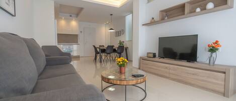 Una sala moderna con un sofá gris, una mesa de centro con un jarrón de flores, un televisor sobre un mueble de madera y una cocina al fondo.