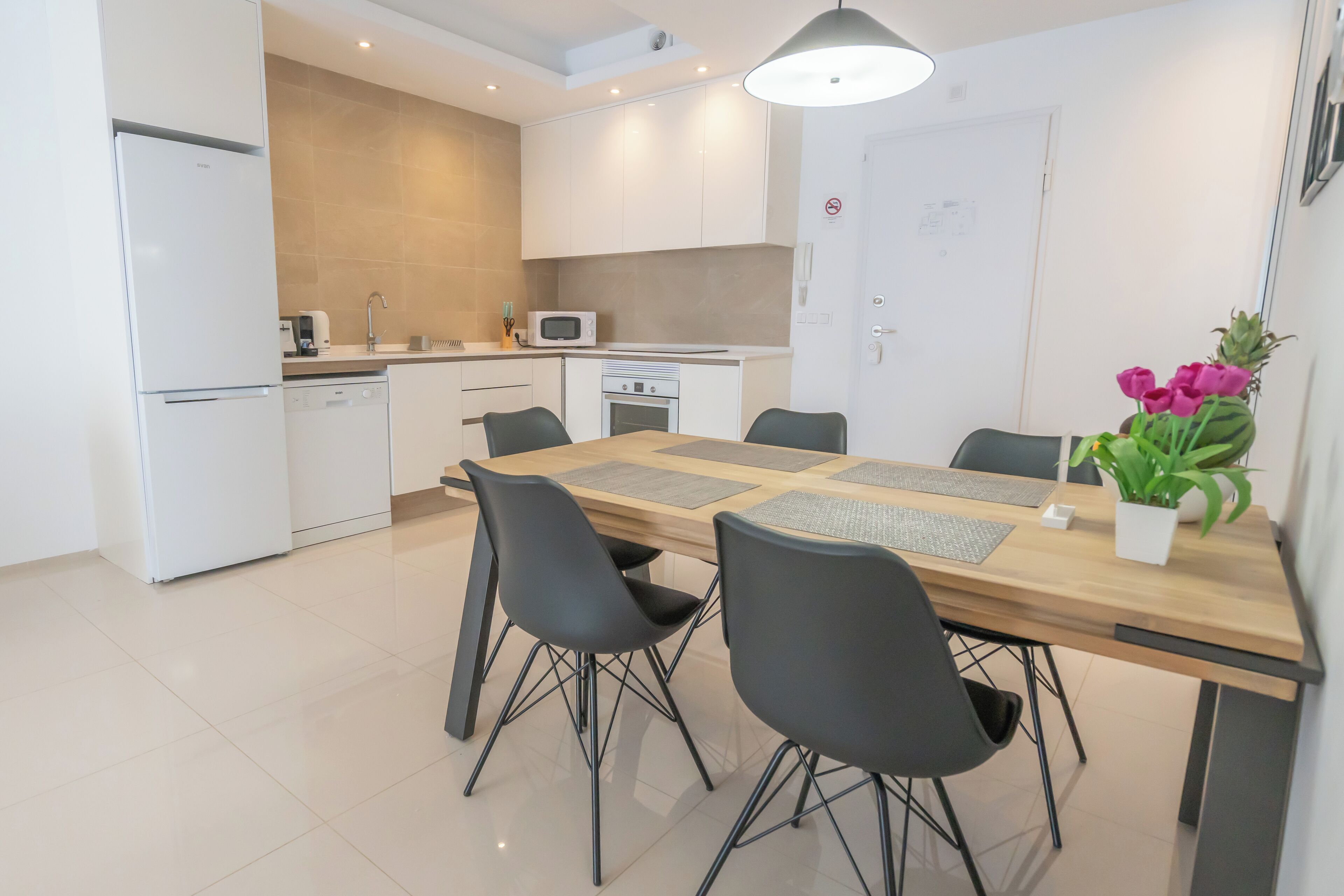 A modern kitchen with white appliances, a dining area with a wooden table and black chairs, and a potted plant on the table.