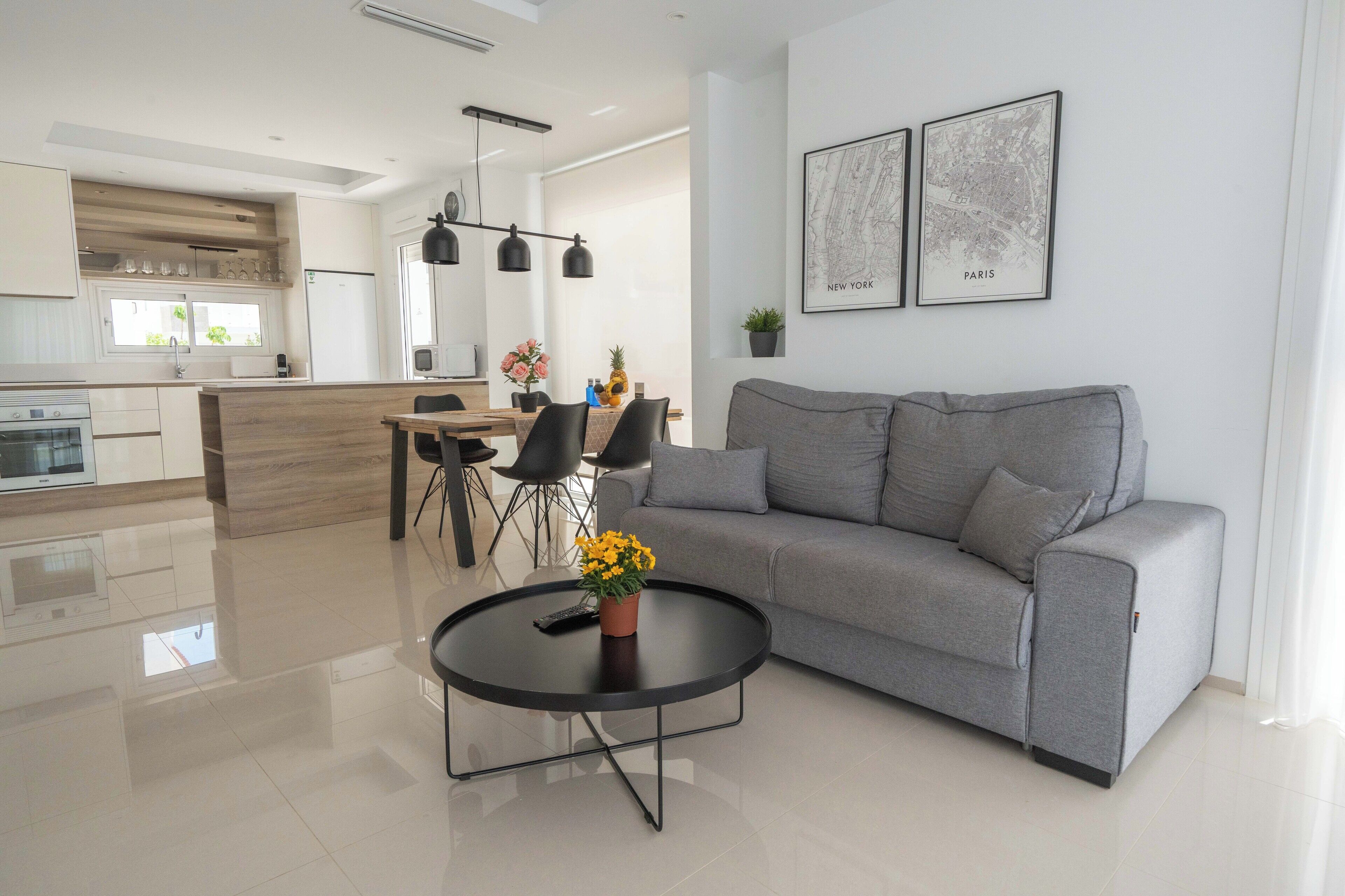 Ein modernes Wohnzimmer mit einem grauen Sofa, einem runden Couchtisch mit Blumenvase und einem Essbereich mit Holztisch und Stühlen.