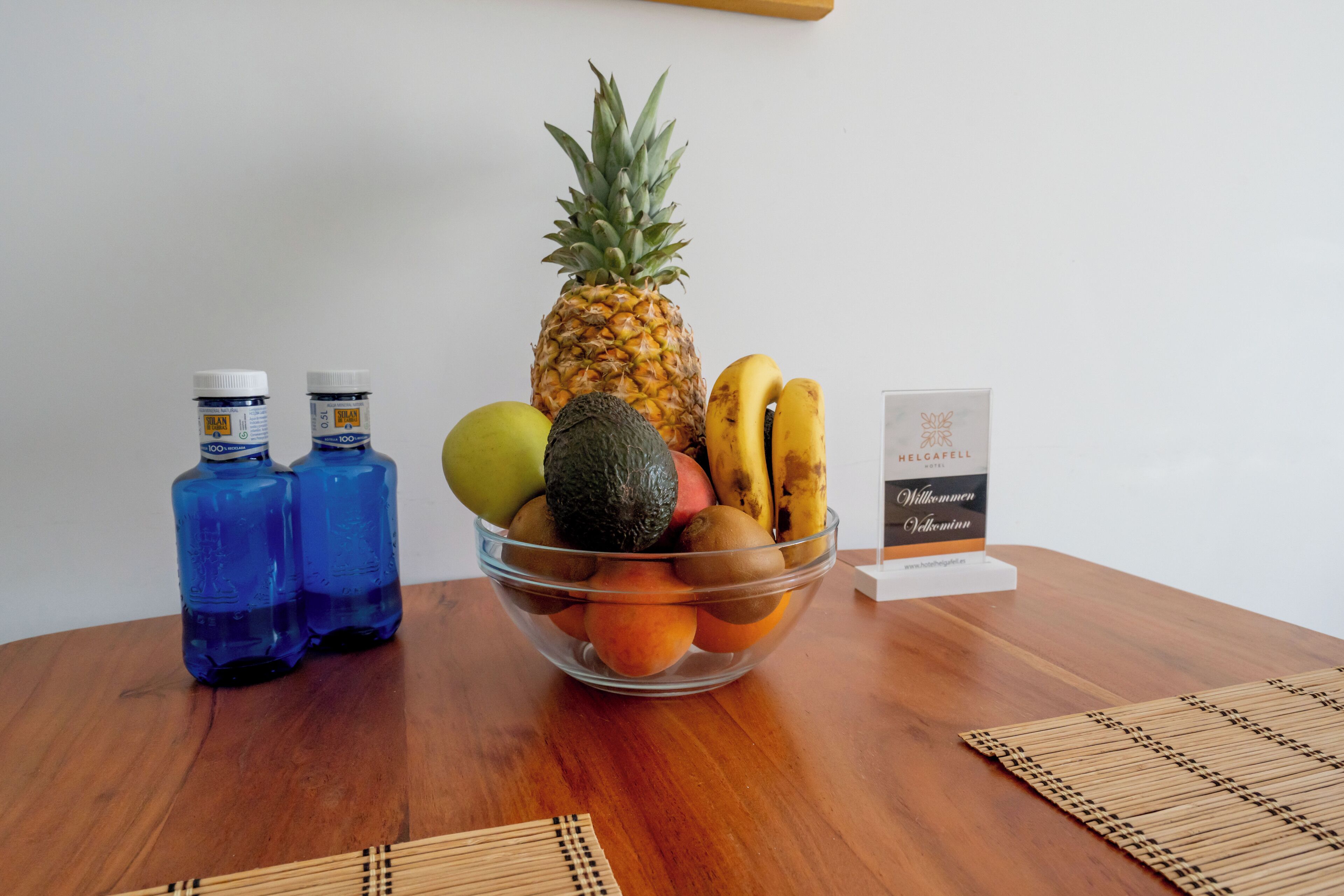 Une table en bois sur laquelle se trouve un panier de fruits contenant un ananas, des bananes, des pommes et des kiwis, ainsi que deux bouteilles d’eau bleues.