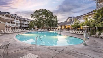 Un espace piscine avec des chaises longues, un grand arbre et un bâtiment doté de balcons.