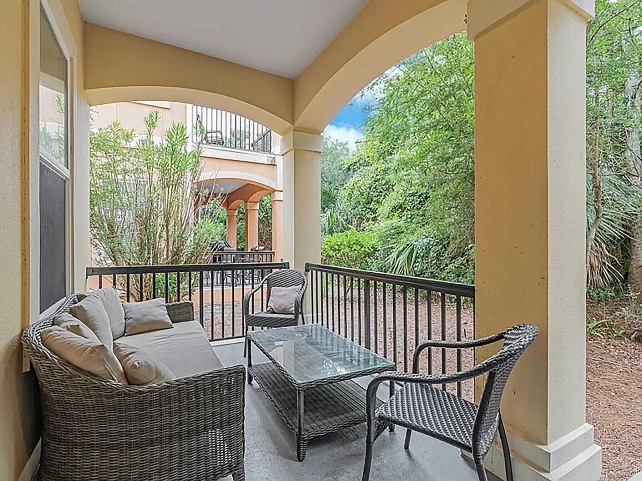 A balcony with wicker furniture, a glass table, and a view of greenery and a building in the background.