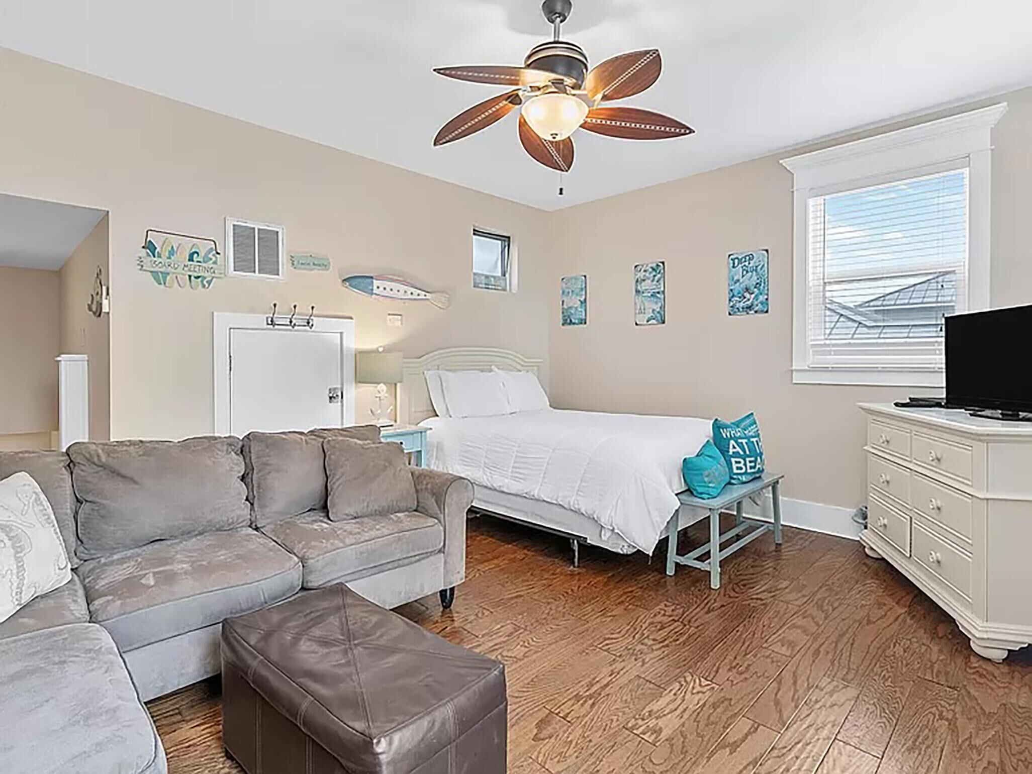 A bedroom with a bed, a gray sofa, a wooden floor, a ceiling fan, and a dresser.