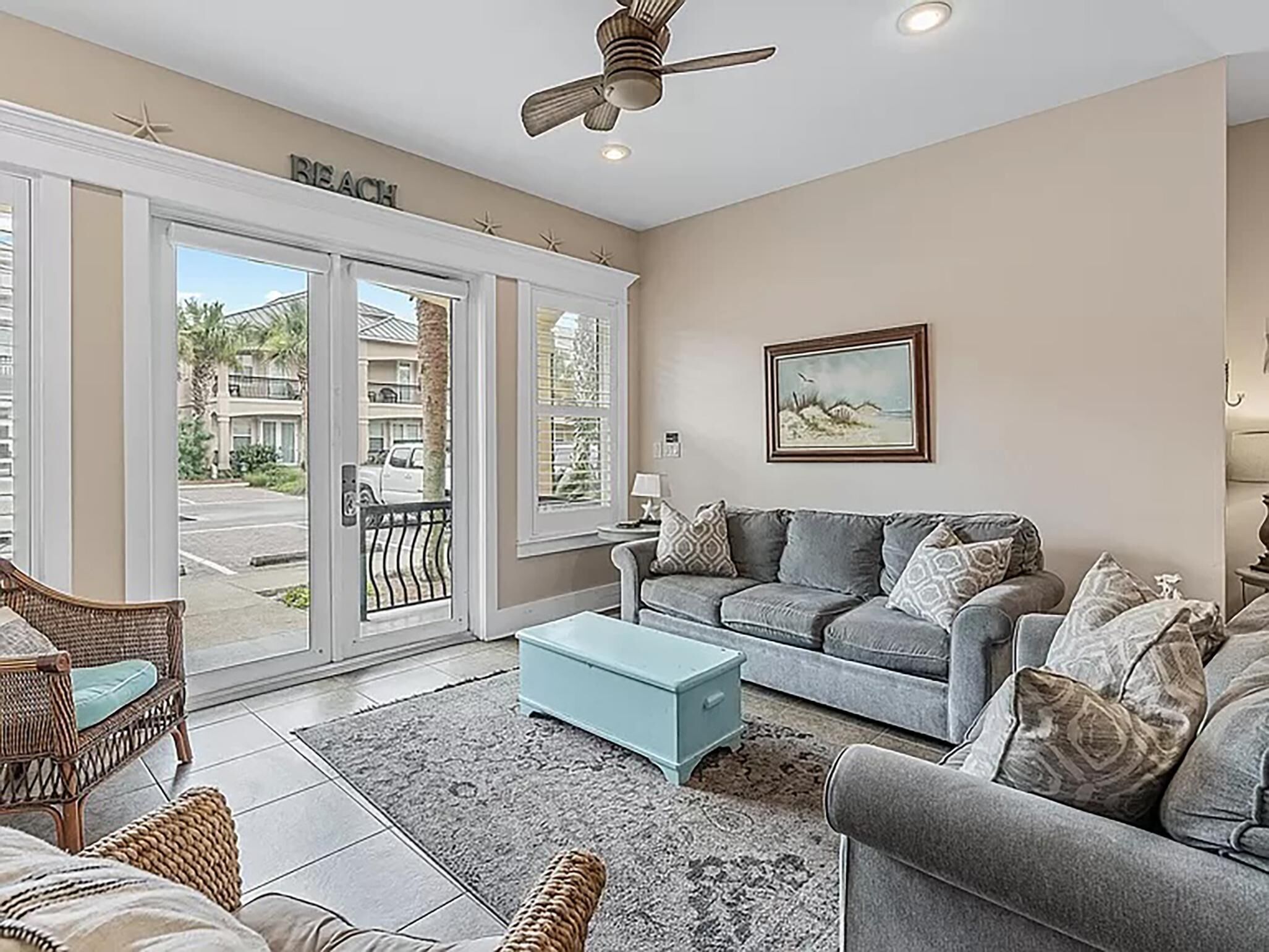A living room with a grey sofa, a wicker chair, a blue ottoman, and a ceiling fan.