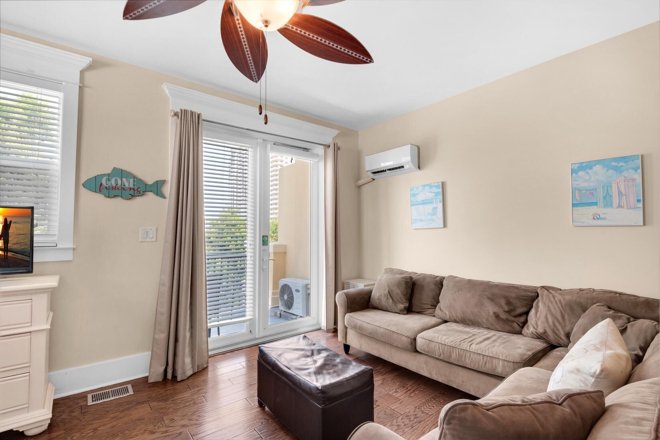 Un salon avec un canapé beige, un pouf foncé, un ventilateur de plafond et une fenêtre avec des stores.