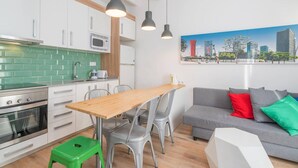 Una cocina moderna con armarios blancos, salpicadero verde y electrodomésticos de acero inoxidable. Hay una mesa de comedor de madera con sillas, un sofá gris con cojines de colores y un cuadro de un paisaje urbano en la pared.