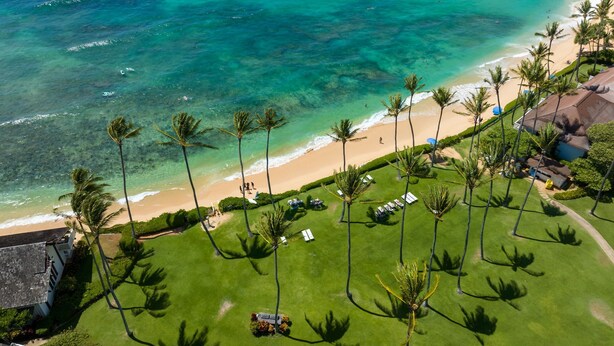 Vista aérea de un resort frente al mar, con césped cuidado, palmeras y una playa de aguas turquesa cristalinas.