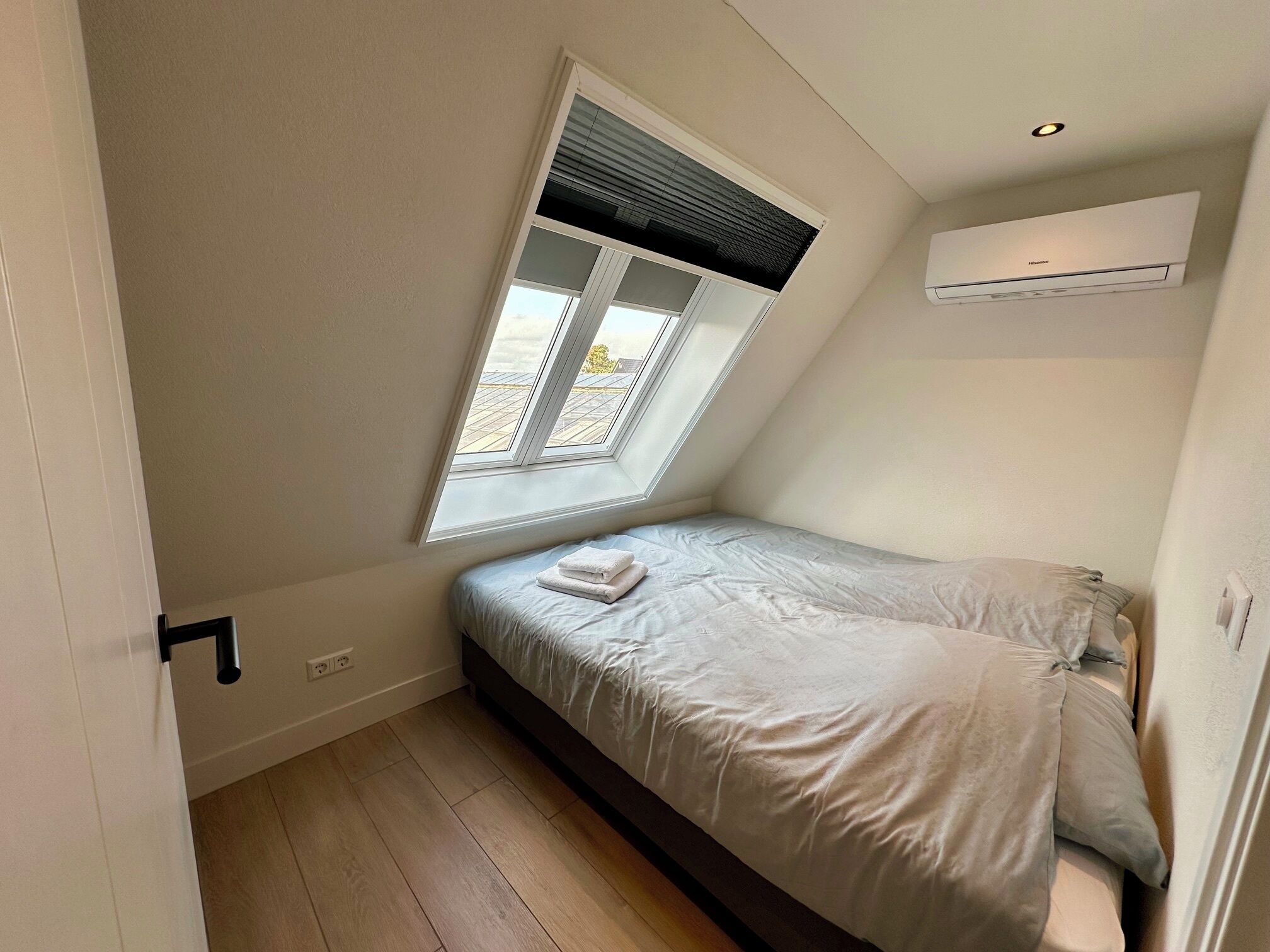 Ein kleines Zimmer mit einem Einzelbett, geneigter Decke, einem Fenster mit Jalousien, einer Wand-Klimaanlage und Holzboden.