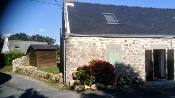 Un bâtiment en pierre doté d’une petite fenêtre et d’une porte, entour é d’un mur de pierre et d’un jardin où poussent des fleurs roses.