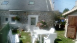 Une maison en pierre avec une porte blanche, un petit jardin avec une table et des chaises, et une voiture garée.
