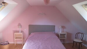 Une chambre avec un plafond en pente, un lit recouvert d’une couverture rose, deux tables de chevet avec des lampes, une chaise et un petit panier posé sur le sol.