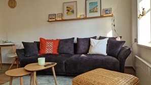 Ein Wohnzimmer mit einem dunkelgrauen Sofa, einem kleinen runden Tisch, einem gewebten Pouf und einem Regal mit gerahmten Bildern.