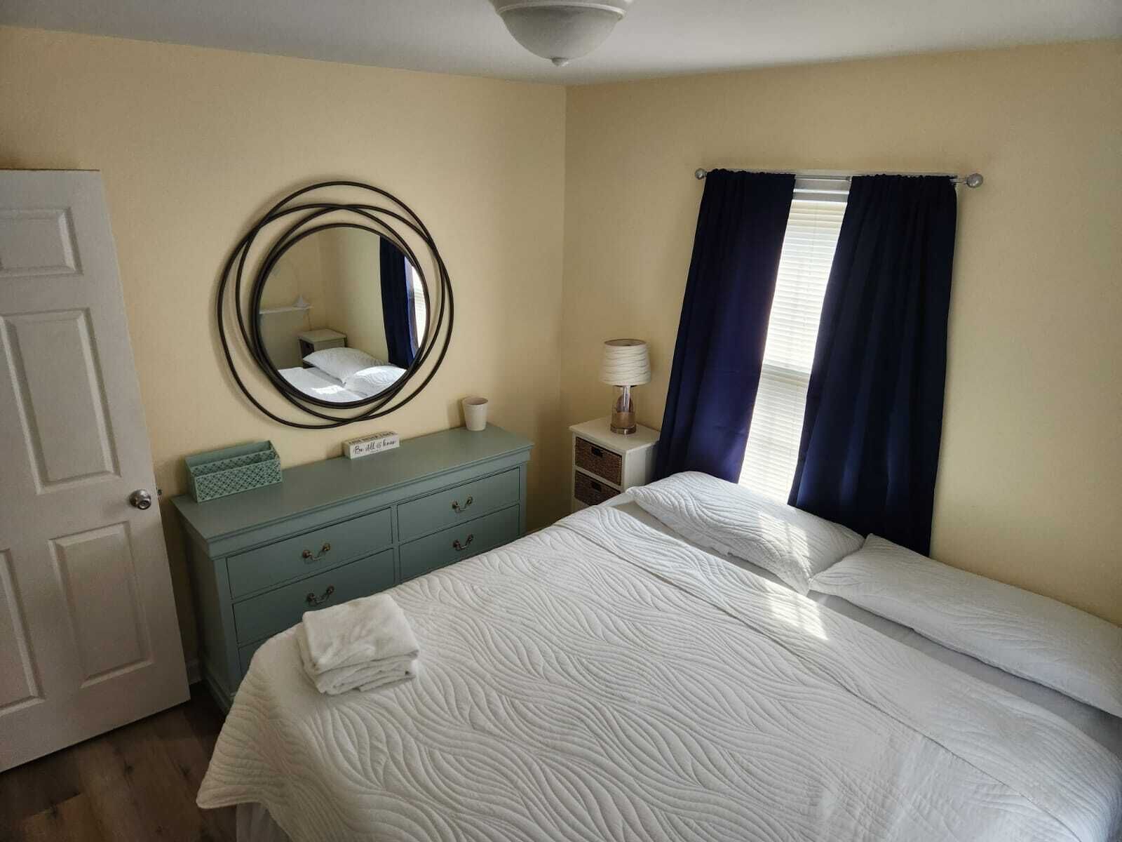 Une chambre à coucher avec un lit, une commode, un miroir, une table de chevet et une fenêtre avec des rideaux.