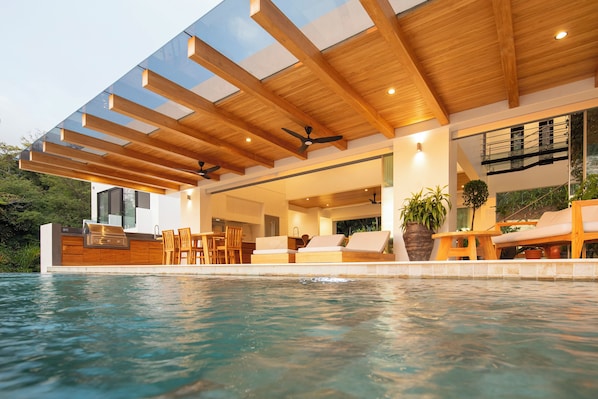 Un área de piscina moderna al aire libre con terraza de madera, asientos y una parrilla integrada.