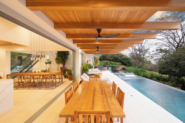 A modern outdoor dining area with a pool, wooden ceiling, and a view of the surrounding garden.