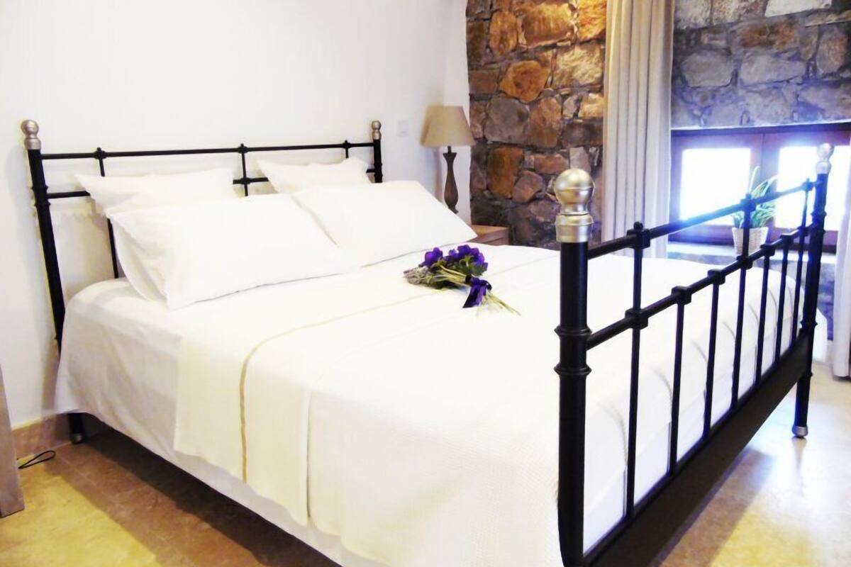A bed with white linens and a bouquet of purple flowers, set against a stone wall and a window with curtains.