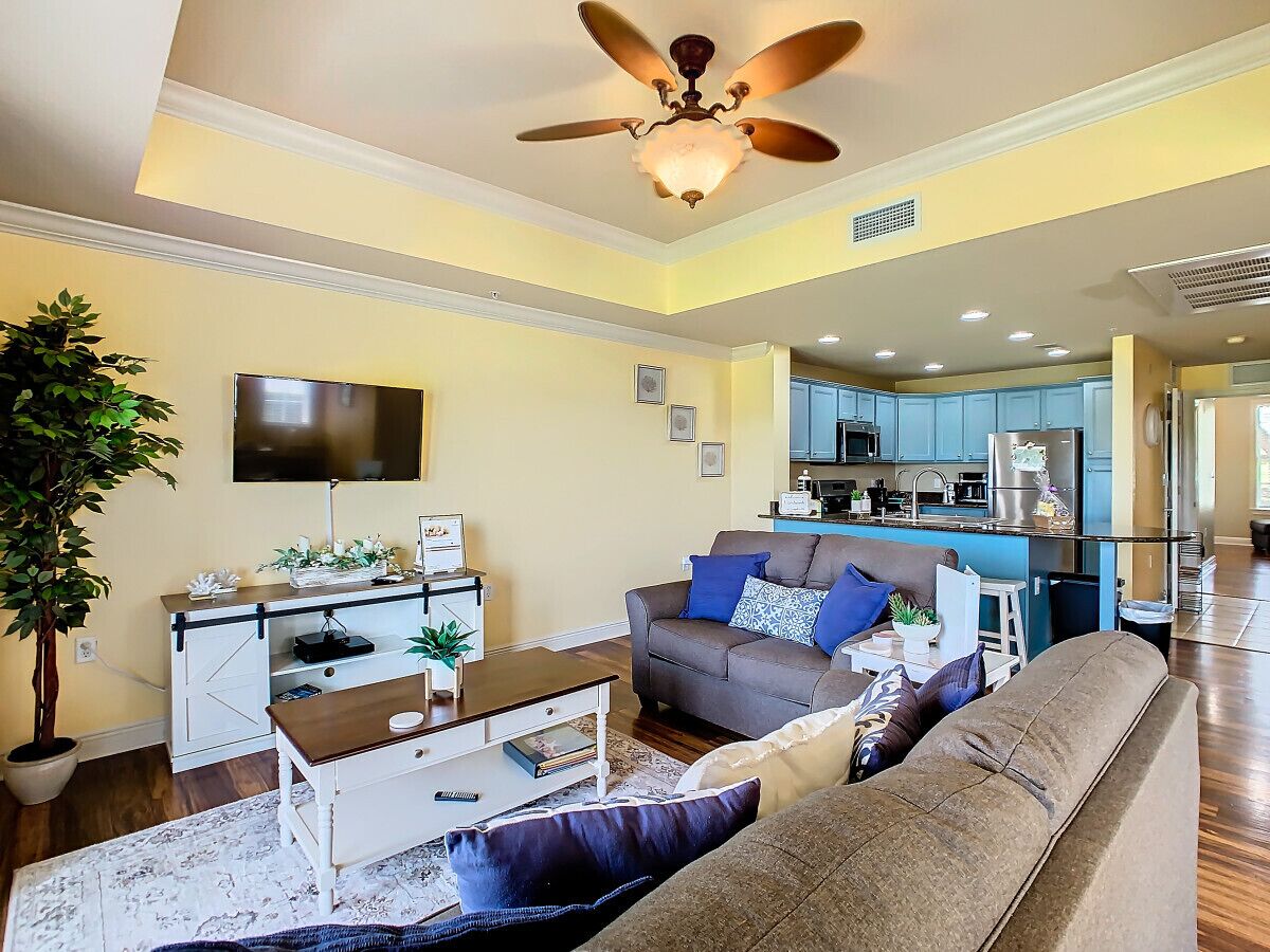 A modern living room with a sofa, coffee table, and TV. There's a kitchen in the background with blue cabinets and a ceiling fan.
