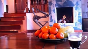 Ein Esstisch mit einer Schale Orangen und einem Glas dunklem Getränk, ein Kamin aus Stein sowie Holzmöbel.