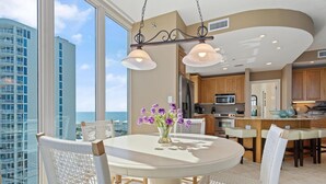 A modern kitchen with a central island, a dining area with a round table and white chairs, and a view of a high-rise building and the ocean. - Lagoon Tower 1002 (Gulf Shores)