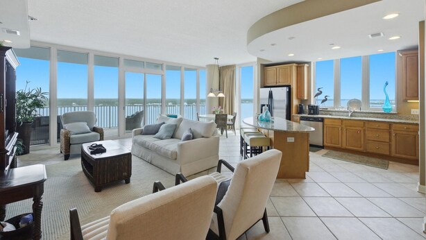 A spacious living area with a sofa, armchairs, and a coffee table. A kitchen with wooden cabinets and a view of the sea is visible. - Lagoon Tower 1002 (Gulf Shores)