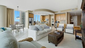 A spacious living area with a sofa, coffee table, and dining table, connected to a kitchen with cabinets and appliances. - Lagoon Tower 1002 (Gulf Shores)