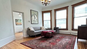 A spacious living room with hardwood flooring, a red patterned area rug, a grey sofa, a brown ottoman, and a chandelier.